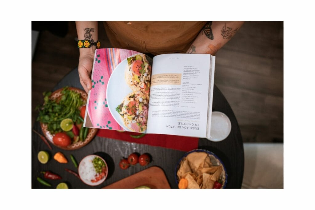 Woman holding cookbook open over a table with vegetables on it.